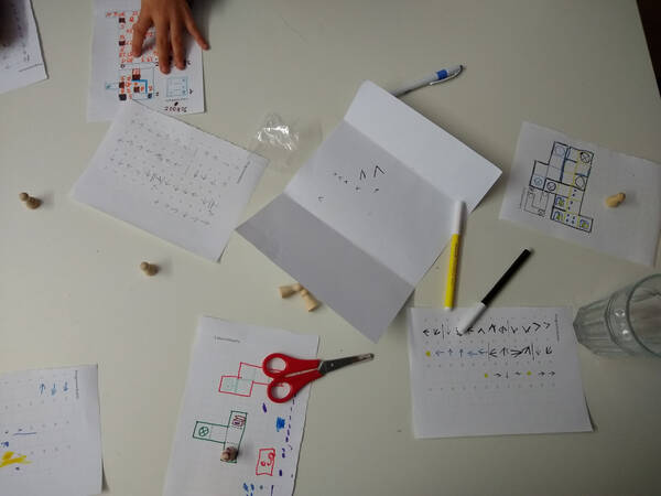 Zettel, Labyrinthe und Programme, liegen auf einem Tisch verteilt; dazwischen Stifte, Spielfiguren und eine Schere.
